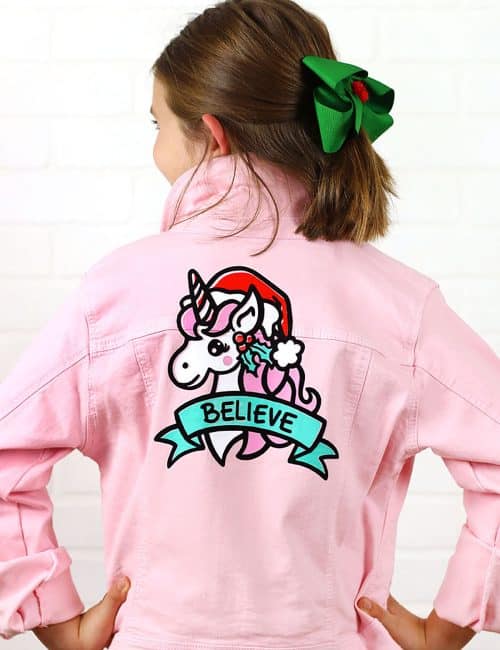 pink denim jacket with christmas unicorn design on back that reads "believe"