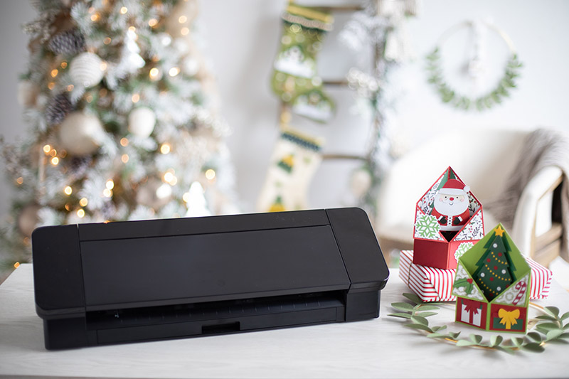 black cameo 4 on a table with paper christmas cards in front of a christmas tree and other christmas decorations