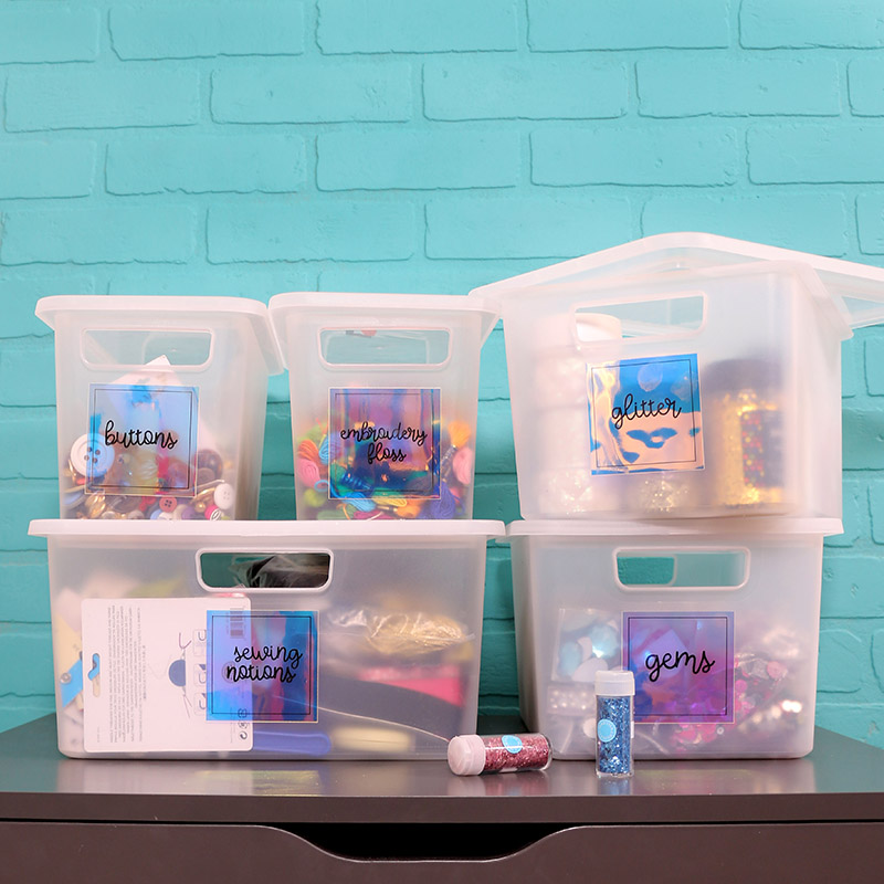 Five clear plastic bins filled with craft supplies sit on a gray table in front of a turquoise brick wall. The bins have iridescent labels on them that read "buttons," "embroidery floss," "sewing notions," "glitter," and "gems." Two small jars of glitter sit on the table in front of the bins.