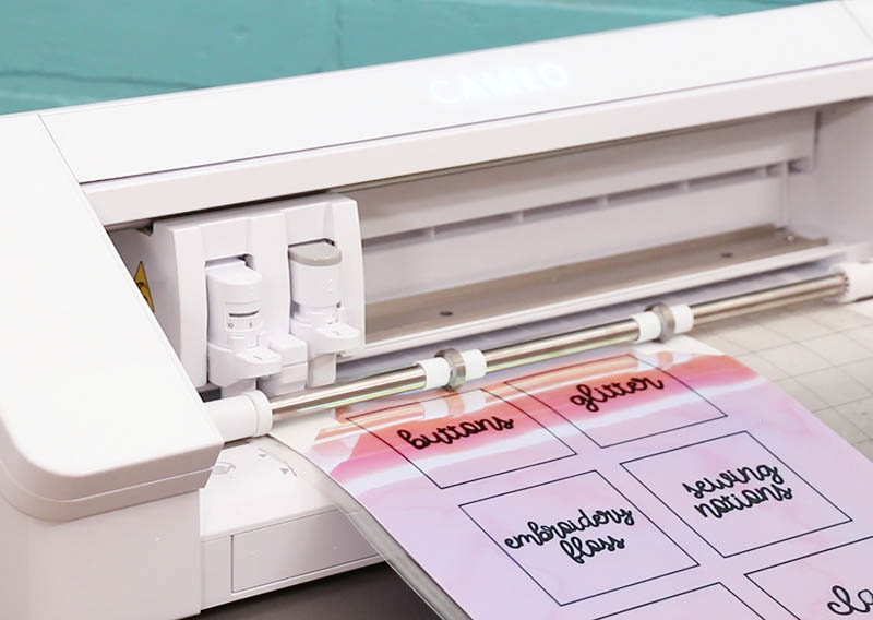 A white Silhouette Cameo 4 machine cuts labels from iridescent sticker paper.