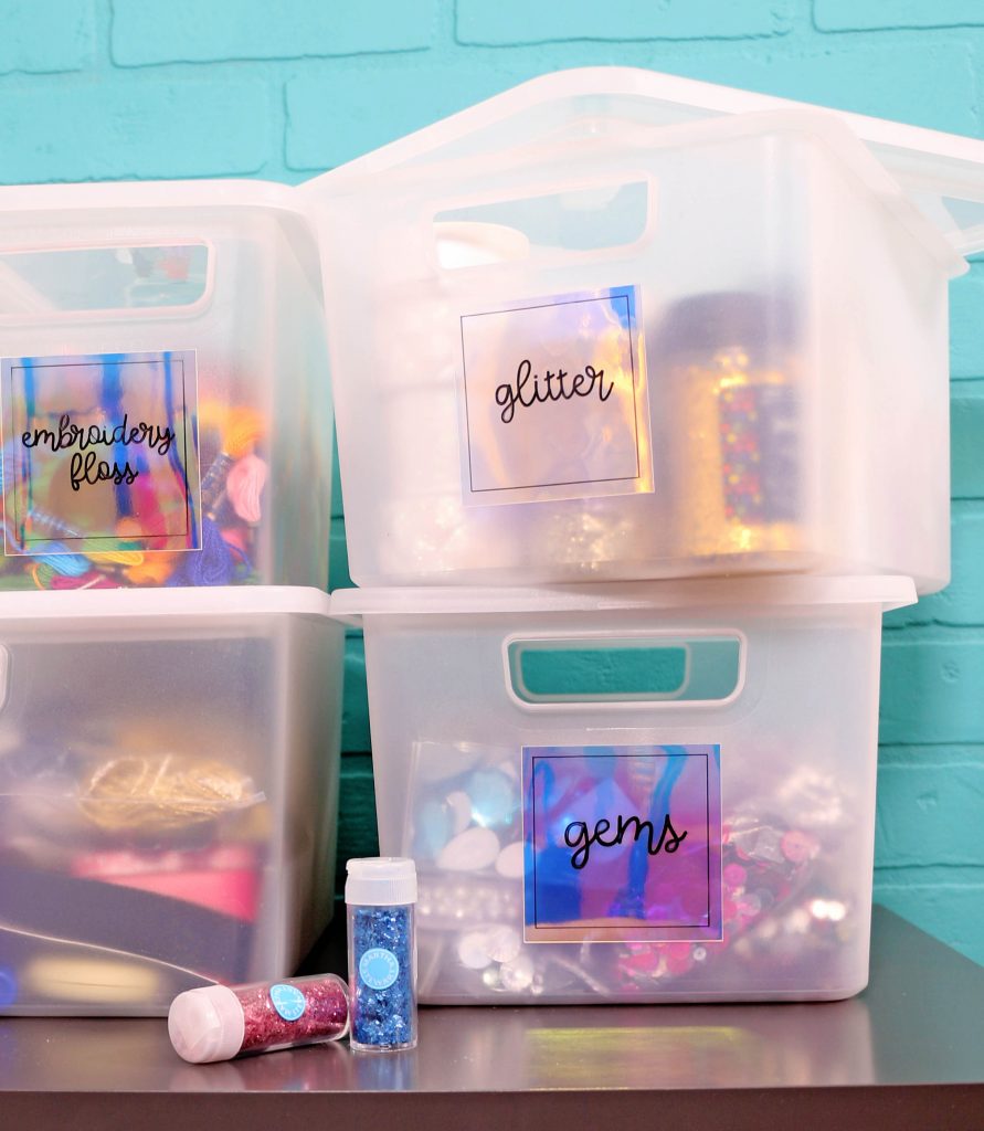 Clear plastic bins filled with craft supplies sit on a gray table in front of a turquoise brick wall. Two small jars of glitter sit on the table in front of the bins.