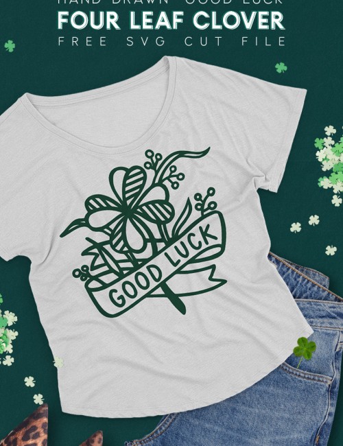 Gray t-shirt on dark green background surrounded by jeans, leopard print shoes, and paper four leaf clover confetti. The t-shirt has a dark green design on it depicting a hand drawn four leaf clover with a ribbon banner that reads "good luck."