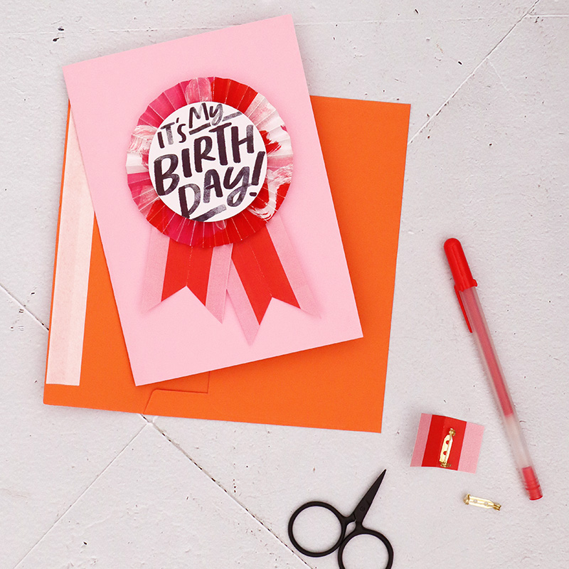 Light pink notecard with pink and orange paper birthday button pinned on the front of the card. An orange envelope, pins, and pen surround the card on a white background.