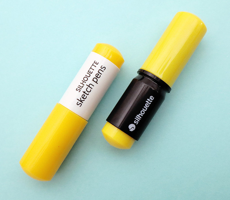 Two yellow Silhouette Sketch Pens - one with a white body and one with a black body - on a light blue blackground.