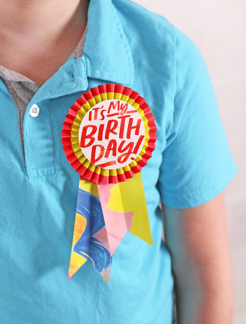 Child wearing light blue polo shirt with paper rosette button pinned on shirt. Button reads it's my birthday!