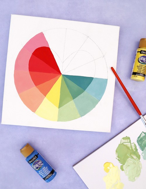 partially painted color wheel art on light purple background surrounded by paints and a paint brush