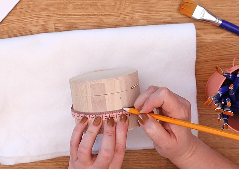 One hand holds a small wooden box with a pink tape measure wrapped around it. Light pencil marks are made every one centimeter.