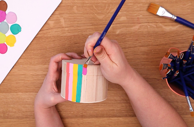 Painting a purple stripe next to other colored stripes on the side of a small wooden box.