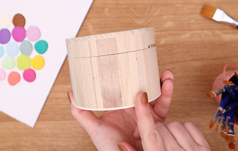 Hands holding a small round wooden box. One finger points out the vertical stripes sketched out on the side of the box with a pencil.