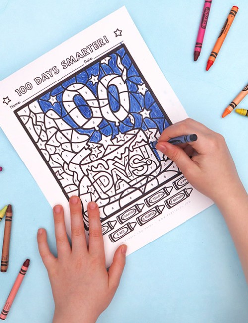 Hands of a child coloring a "100 Days Smarter" color by number coloring page. The picture the child is coloring shows number balloons and a cake that read "100 Days." Crayons are scattered around the light blue background.