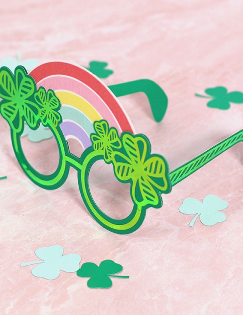 Green paper St. Patrick's Day glasses with holographic four leaf clover designs and a paper rainbow along the top of the glasses sitting on a light pink background.