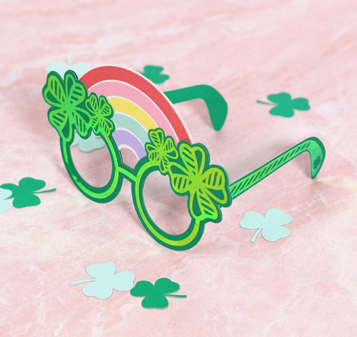 Green paper St. Patrick's Day glasses with holographic four leaf clover designs and a paper rainbow along the top of the glasses sitting on a light pink background.