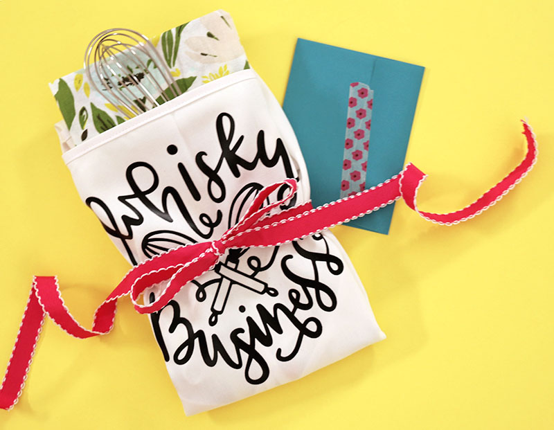 White whisky business apron folded up with whisk inside it and tied up with a ribbon on yellow background with notecard tucked underneath the gift