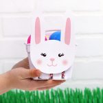 Hands holding a paper berry basket filled with eggs with a white and pink paper bunny face glued on the front