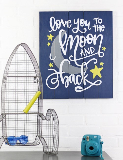 Painted wooden sign that reads "love you to the moon and back" in white script lettering on a dark blue background with a gray moon and yellow stars. The sign hangs on a white wall with a gray spaceship shaped wire shelf and bright blue camera sitting on a gray cabinet in the foreground.