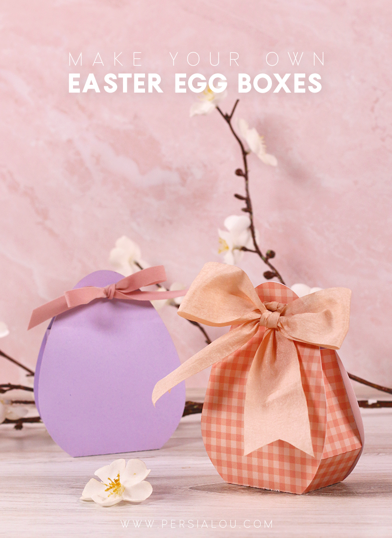 Two paper egg shaped boxes tied closed with ribbons sit on a light wood surface surrounded by cherry blossoms