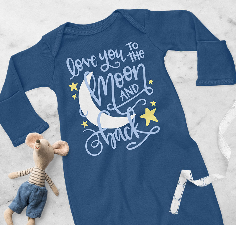 Dark blue baby layette with hand lettered "to the moon and back" design on the front laying on white marble background with a soft mouse toy