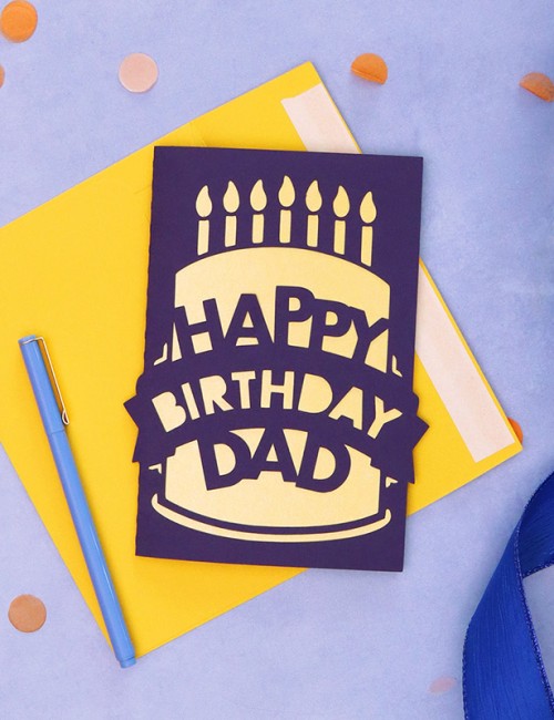 Dark purple and yellow greeting card sitting on top of yellow envelope and surrounded by confetti and curled ribbon. The card has a papercut design on the front depicting a birthday cake that reads 'Happy Birthday Dad'