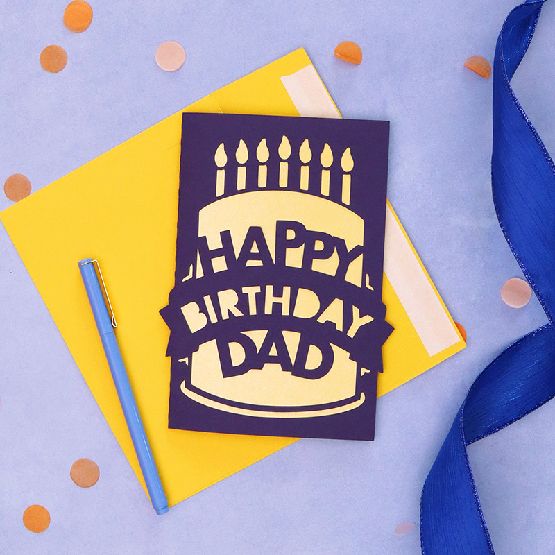 Dark purple and yellow greeting card sitting on top of yellow envelope and surrounded by confetti and curled ribbon. The card has a papercut design on the front depicting a birthday cake that reads 'Happy Birthday Dad'