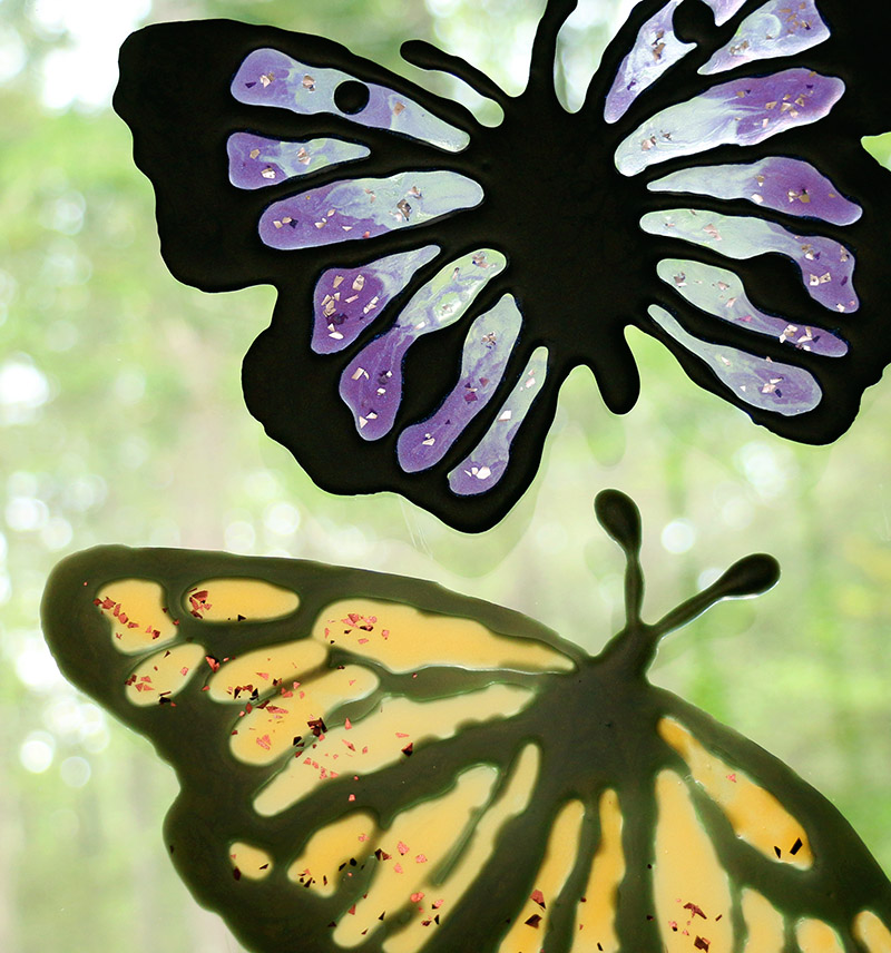 Two butterfly shaped DIY window clings in shades of translucent purple and amber with glitter flakes on a window with foliage in the background.
