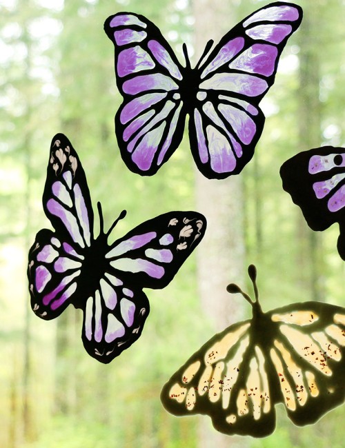 Three butterfly shaped DIY window clings in shades of translucent purple and amber on a window with foliage in the background.