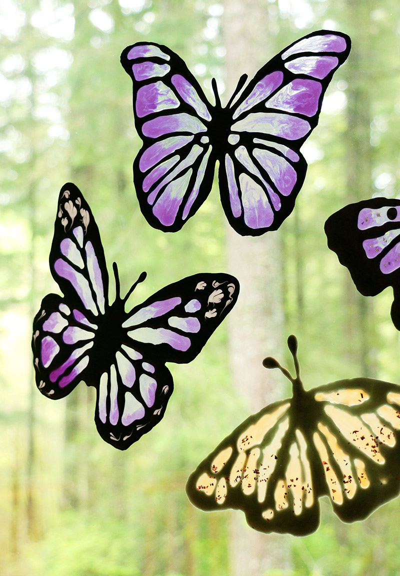 Three butterfly shaped DIY window clings in shades of translucent purple and amber on a window with foliage in the background.