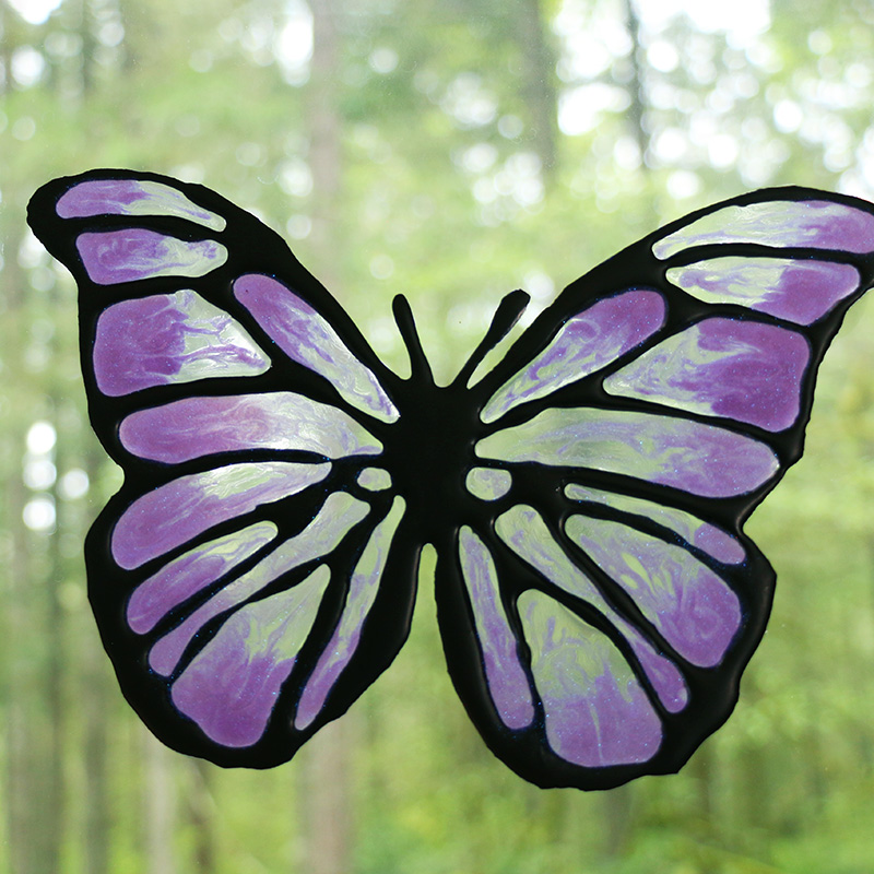 Black, clear, and purple butterfly shaped diy window cling on a window with green foliage in the background.